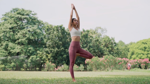 Mujer joven asiática de pie sobre la hierba practicando yoga en la posición de pose de árbol en el parque de la ciudad con el fondo de grandes árboles. Retrovisor de mujer practicando yoga al aire libre en un día soleado.