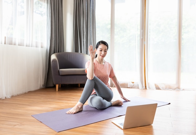 Mujer joven asiática practicando yoga en la estera mientras está sentado frente a su computadora portátil en casa.