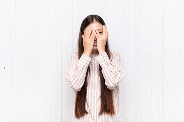 Mujer joven asiática que cubre la cara con las manos, mirando entre los dedos con expresión de sorpresa y mirando hacia un lado