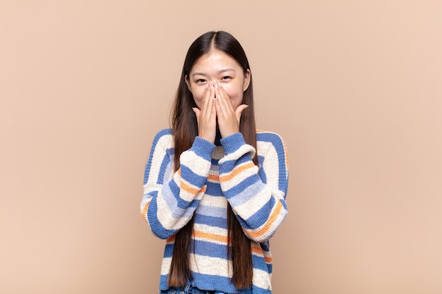 Mujer joven asiática que parece feliz, alegre, afortunada y sorprendida cubriendo la boca con ambas manos