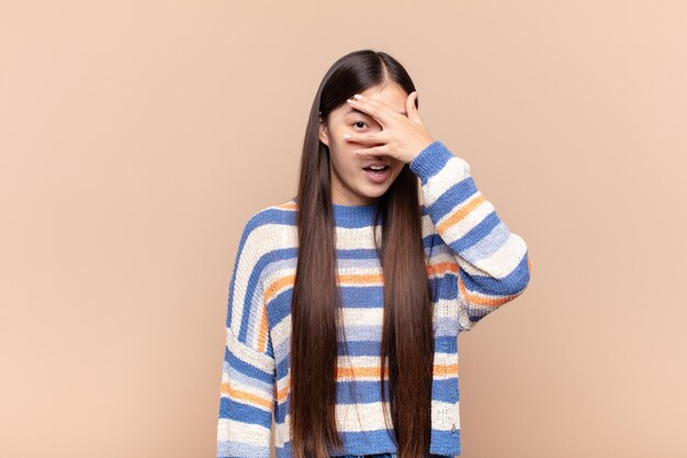 Mujer joven asiática que parece sorprendida, asustada o aterrorizada, cubriendo la cara con la mano y mirando entre los dedos