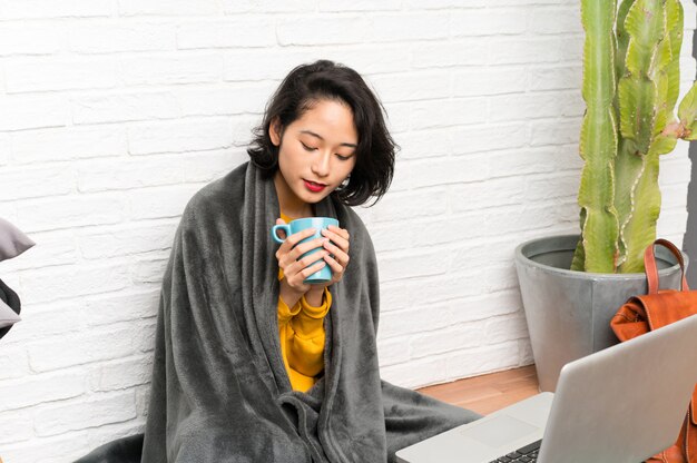 Mujer joven asiática que se sienta en el piso que sostiene una taza de café