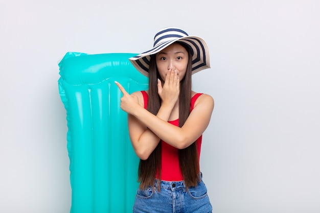 Mujer joven asiática que se siente feliz, conmocionada y sorprendida, cubriendo la boca con la mano y apuntando al espacio lateral de la copia. concepto de verano