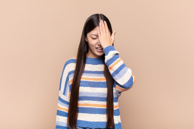 Mujer joven asiática riendo y golpeándose la frente como diciendo ¡oh! Lo olvidé o fue un error estúpido