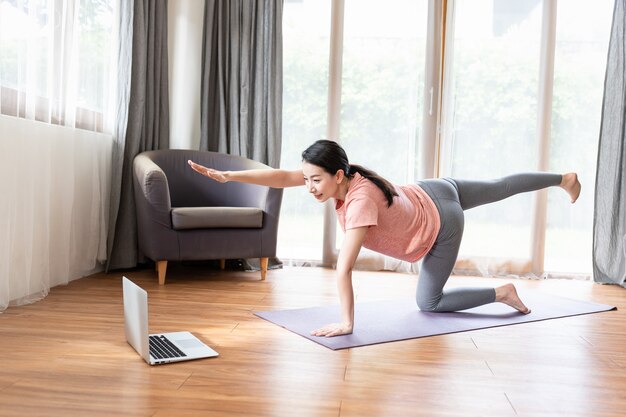 Mujer joven asiática en ropa deportiva haciendo ejercicio o practicando yoga en la estera frente a su computadora portátil en casa y estirando sus manos y piernas.