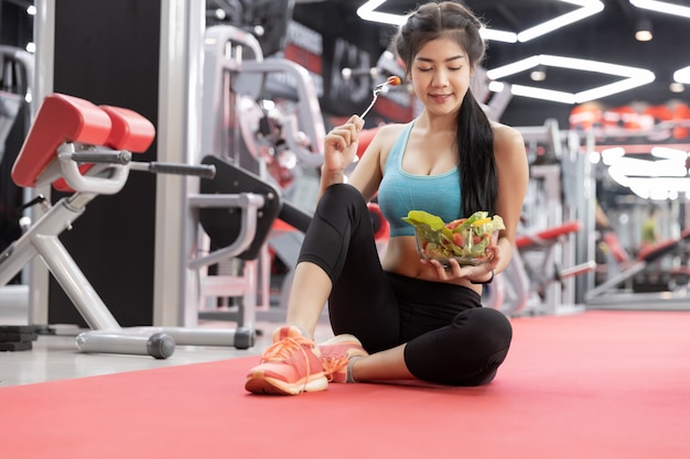 Mujer joven asiática sana que come la ensalada verde de las verduras en el gimnasio.