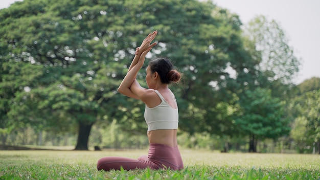 Mujer joven asiática sentada en la hierba practicando yoga en la posición de pose de vaca en el parque de la ciudad con el fondo de grandes árboles. Retrovisor de mujer practicando yoga al aire libre en un día soleado.