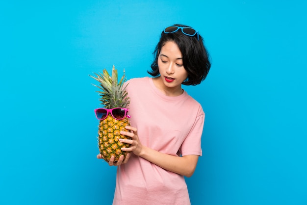 Mujer joven asiática sobre el azul aislado que sostiene una piña con las gafas de sol