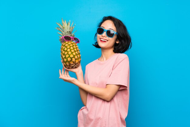 Mujer joven asiática sobre el azul aislado que sostiene una piña con las gafas de sol