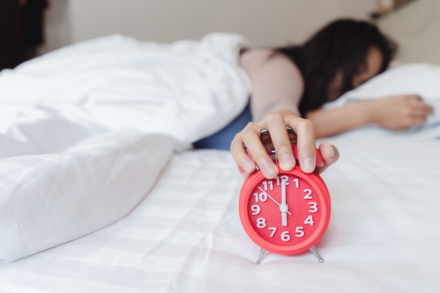 Una mujer joven asiática soñolienta que sostiene un despertadorPara despertarse para dormir