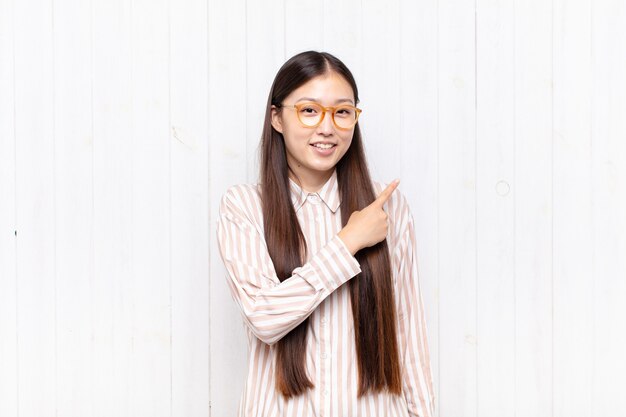 Mujer joven asiática sonriendo alegremente, sintiéndose feliz y apuntando hacia un lado y hacia arriba