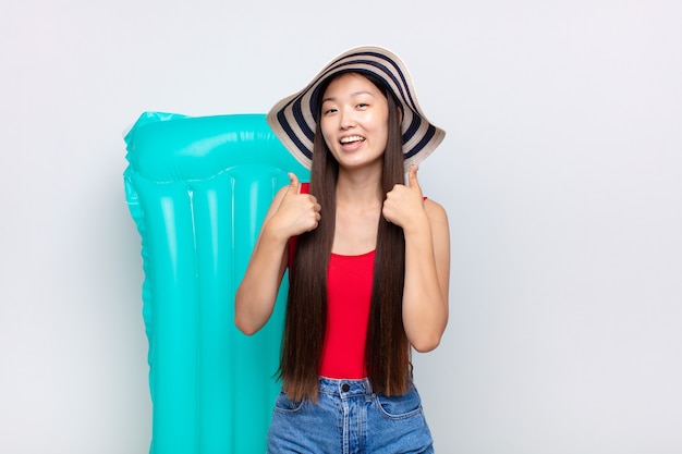 Mujer joven asiática sonriendo ampliamente con aspecto feliz, positivo, seguro y exitoso, con ambos pulgares hacia arriba. concepto de verano