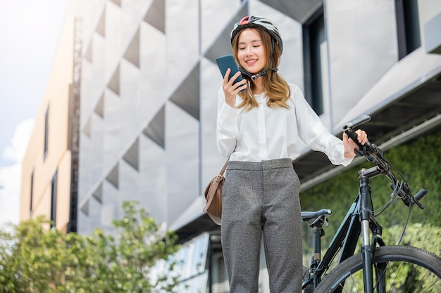 Mujer joven asiática sonriente con casco sostiene una conversación de teléfono inteligente móvil con el negocio durante el trabajo de oficina en la calle con bicicleta, respetuosa con el medio ambiente, mujer de negocios de estilo de vida que viaja afuera por la mañana