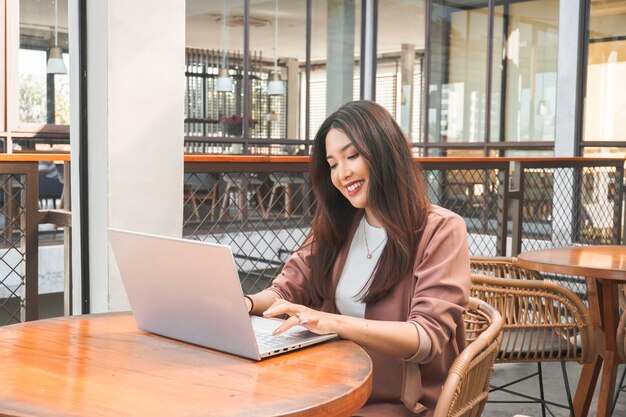Mujer joven asiática sonriente trabajando remotamente desde una cafetería usando su computadora portátil empleada preparándose para una entrevista de videollamada en línea