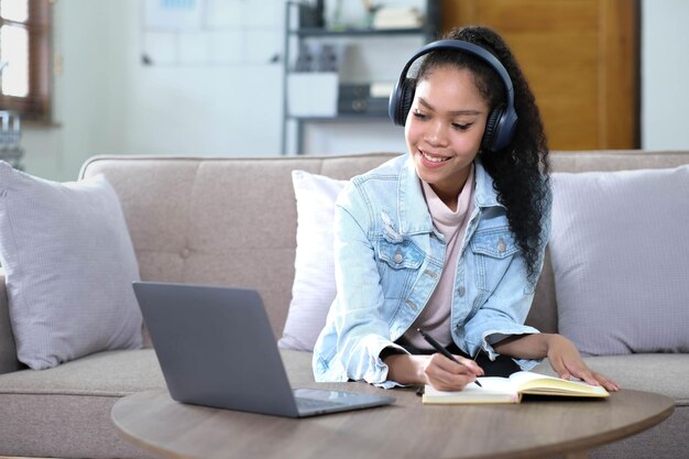 Mujer joven asiática sonriente usando auriculares mirando la pantalla de la computadora portátil escuchando y aprendiendo cursos en línea