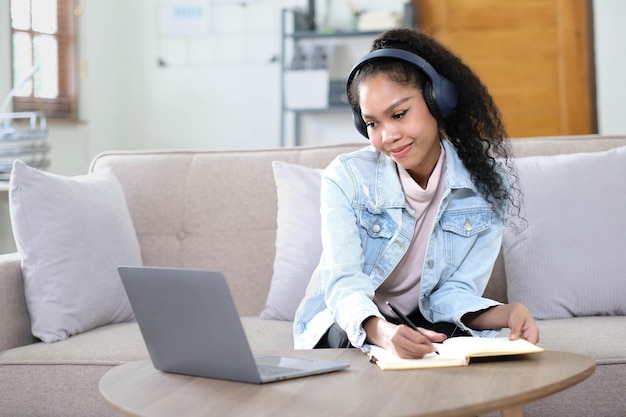 Mujer joven asiática sonriente usando auriculares mirando la pantalla de la computadora portátil escuchando y aprendiendo cursos en línea