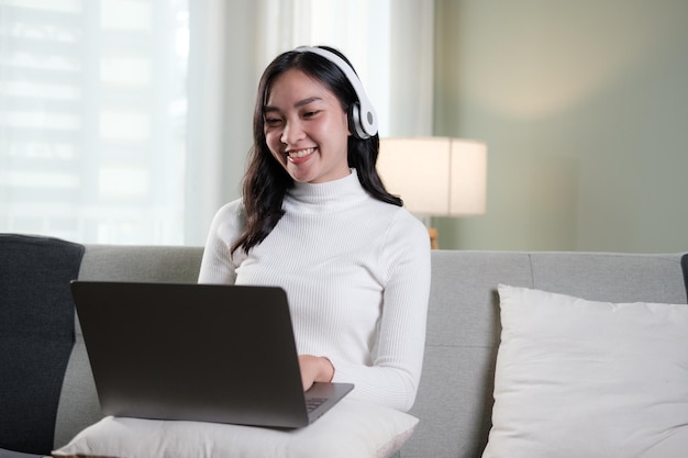 Mujer joven asiática sonriente usando auriculares mirando la pantalla de la computadora portátil escuchando y aprendiendo cursos en línea