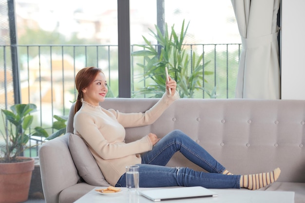 Mujer joven asiática tiene video chat con amigos por teléfono inteligente en la sala de estar en casa