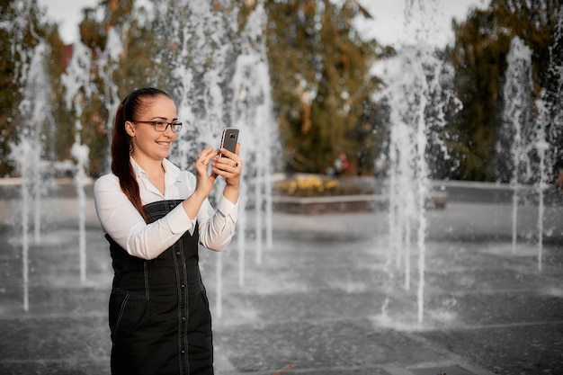 mujer joven aspecto caucásico en gafas ópticas en el parque cerca de la fuente con el teléfono inteligente en la mano.