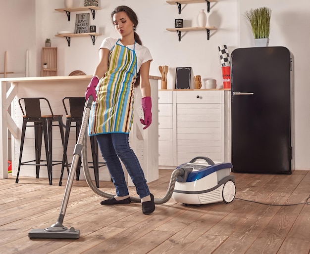 Mujer joven con aspiradora en el piso de la cocina de casa, haciendo tareas de limpieza y quehaceres, interior meticuloso.