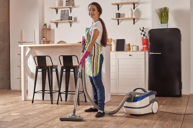 Mujer joven con aspiradora en el piso de la cocina casera haciendo tareas de limpieza y quehaceres meticuloso ...