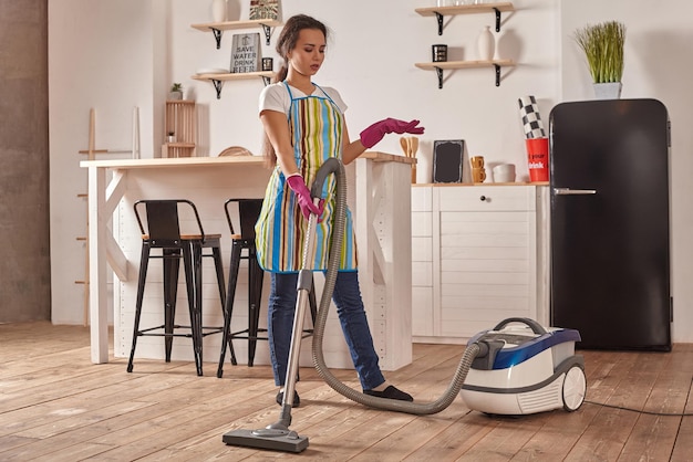 Mujer joven con aspiradora en el piso de la cocina casera haciendo tareas de limpieza y quehaceres meticuloso ...