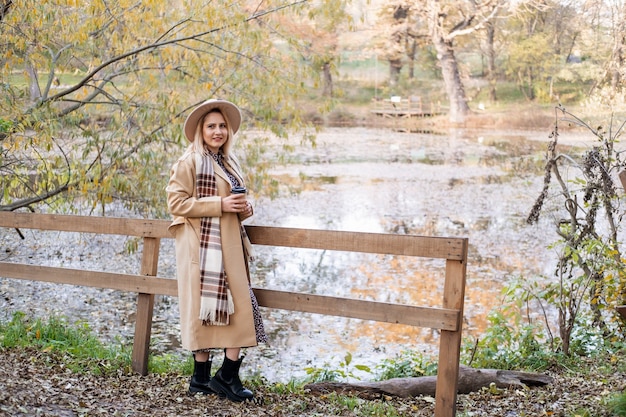 Mujer joven atractiva con abrigo beige y sombrero bebe café junto al lago en el parque de otoño en otoño.