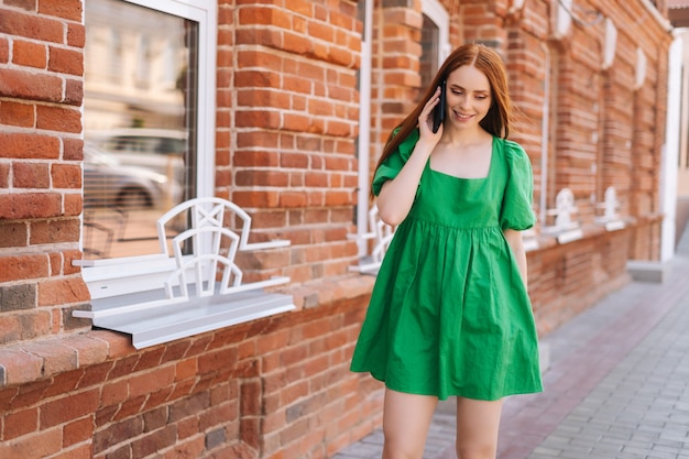 Mujer joven atractiva alegre hablando por teléfono móvil caminando por las calles de la ciudad en un día soleado de verano. Señora sonriente feliz en vestido verde con smartphone al aire libre, fondo borroso, enfoque selectivo.