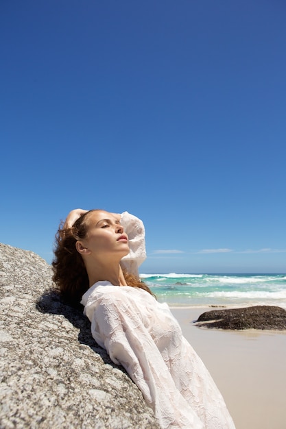 Foto mujer joven atractiva apoyándose en la roca en la playa