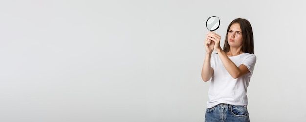 Mujer joven atractiva de aspecto serio que busca algo mirando a través de una lupa blanca
