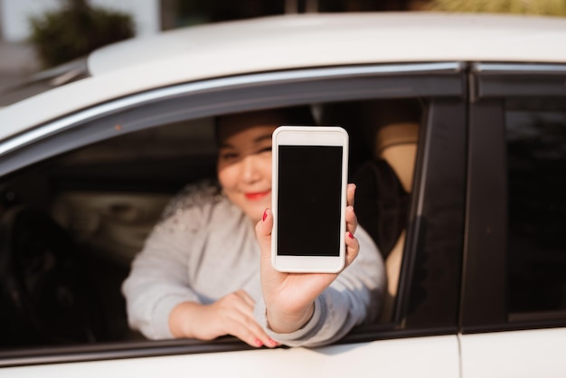 Foto mujer joven atractiva en auto muestra smartphone con pantalla en blanco.