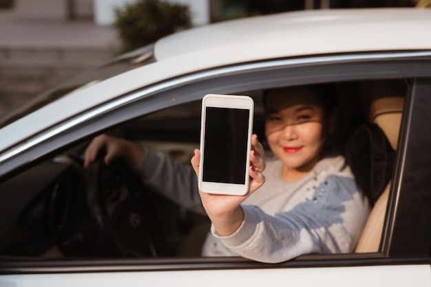 Mujer joven atractiva en auto muestra smartphone con pantalla en blanco.