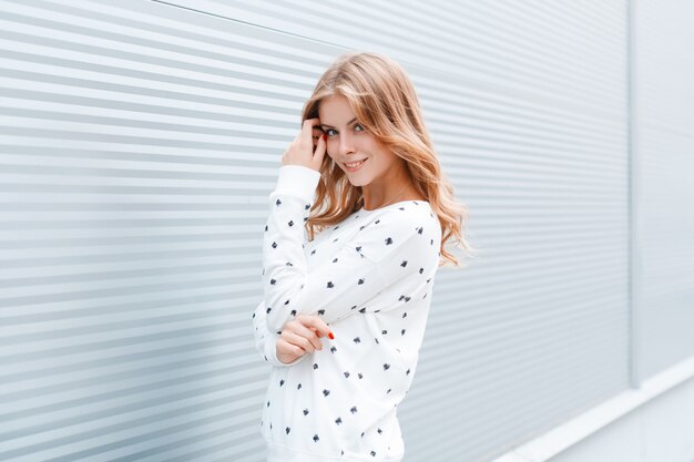 Mujer joven atractiva bastante feliz en elegante pulóver blanco posa al aire libre en la ciudad cerca de la pared de metal moderno en un día soleado