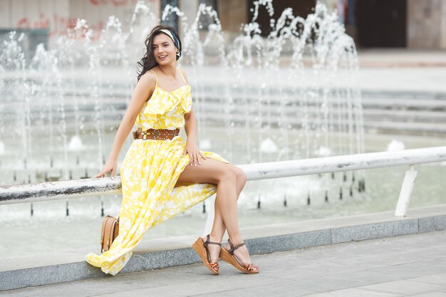 Mujer joven atractiva cerca de la fuente. Hermosa mujer al aire libre. Señora en retrato de vestido amarillo.