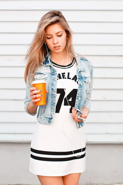 Mujer joven atractiva con una chaqueta de mezclilla de moda con un elegante vestido deportivo con auriculares blancos en los oídos se encuentra cerca de una pared de madera. Chica sexy bebe café caliente y escucha música.