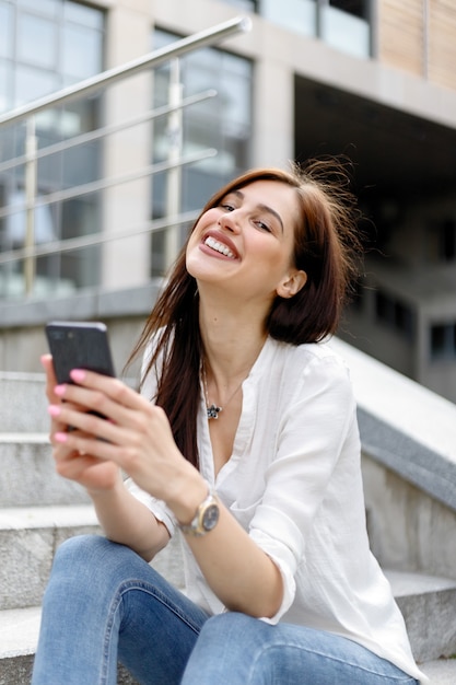 Mujer joven atractiva en la ciudad. Empresaria caucásica con smartphone con la mano. Retrato de mujer de negocios con estilo en ropa de moda con teléfono cerca del edificio de oficinas.