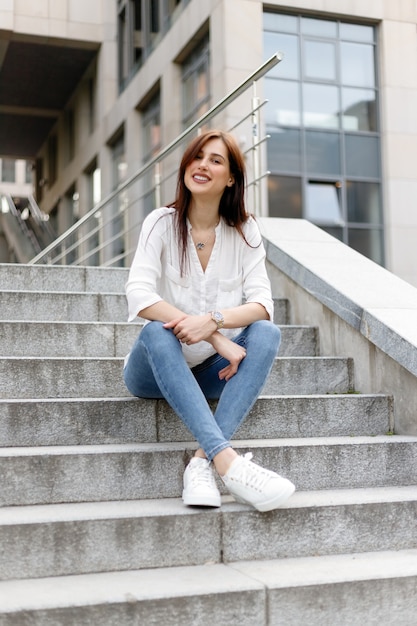 Mujer joven atractiva en la ciudad. Señora de negocios de pie cerca del centro de negocios. Mujer de negocios cansada con camisa blanca en jeans azul sentado relajarse en la calle cerca del edificio de cristal del centro de la ciudad