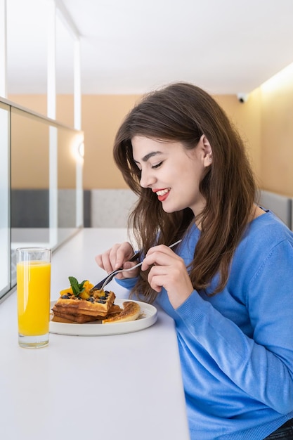 Mujer joven atractiva come gofres belgas y jugo para el desayuno