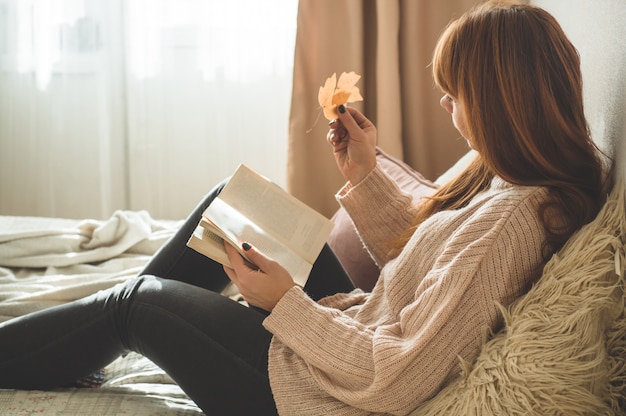 Mujer joven atractiva está leyendo un libro y sostiene una hoja de otoño en su mano