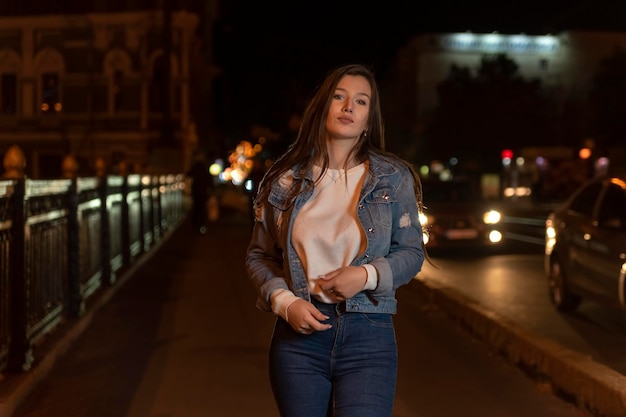 Foto mujer joven atractiva con estilo en el fondo de la ciudad de la noche de la chaqueta del dril de algodón retrato de la estudiante que camina en la calle de la noche