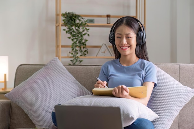 Mujer joven atractiva y feliz escuchando música en auriculares con placer en la sala de estar acostada en el sofá usando portátil y descansando
