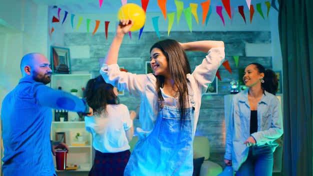 Foto mujer joven atractiva de fiesta con sus amigos mientras sostiene un globo. fiesta universitaria salvaje con una sala llena de luces de neón, bola de discoteca y alcohol