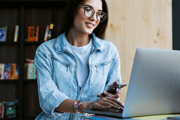 Mujer joven atractiva con gafas se encuentra cerca de la ventana, tiene el teléfono inteligente en la mano y trabaja en la computadora portátil.