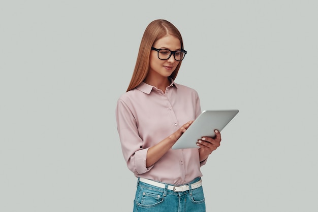 Mujer joven atractiva en gafas con tableta digital y sonriendo mientras está de pie contra el fondo gris