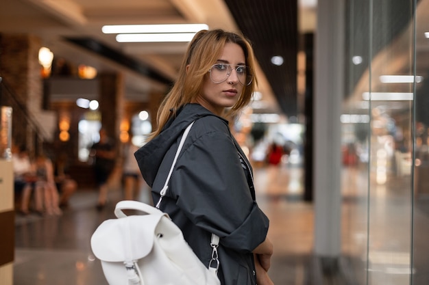 Mujer joven atractiva con un impermeable vintage en gafas de moda con una elegante mochila de cuero blanco es camina cerca de una pared de cristal moderna en un centro comercial. Hermosa chica. Estilo juvenil.