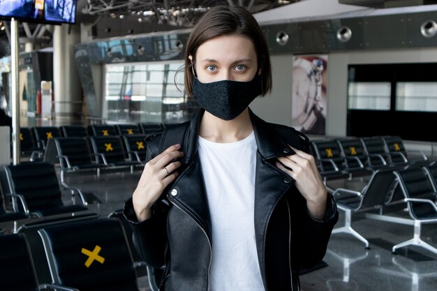 Mujer joven atractiva con una máscara protectora negra con una mochila en la terminal del aeropuerto internacional. Viajar durante la pandemia de coronavirus.