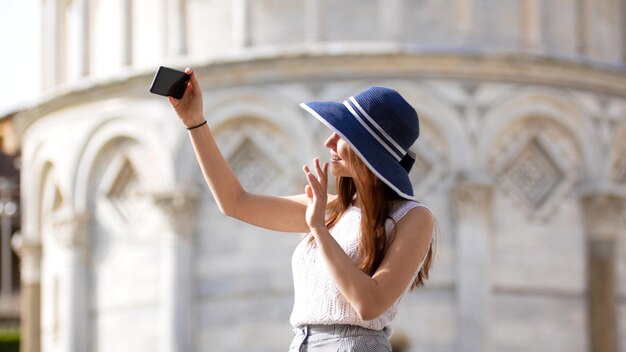 Una mujer joven y atractiva parada bajo la torre de Pisa se toma una selfie y saluda con la mano