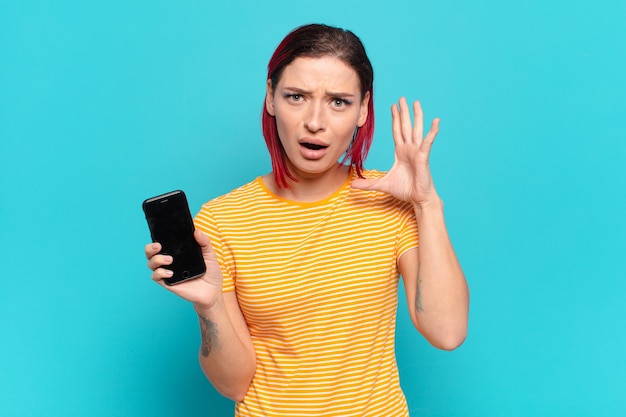 Mujer joven atractiva pelirroja gritando con las manos en el aire, sintiéndose furiosa, frustrada, estresada y molesta y mostrando su celular