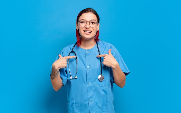 Mujer joven atractiva pelirroja que se siente feliz, sorprendida y orgullosa, señalando a sí misma con una mirada emocionada y asombrada. concepto de enfermera del hospital