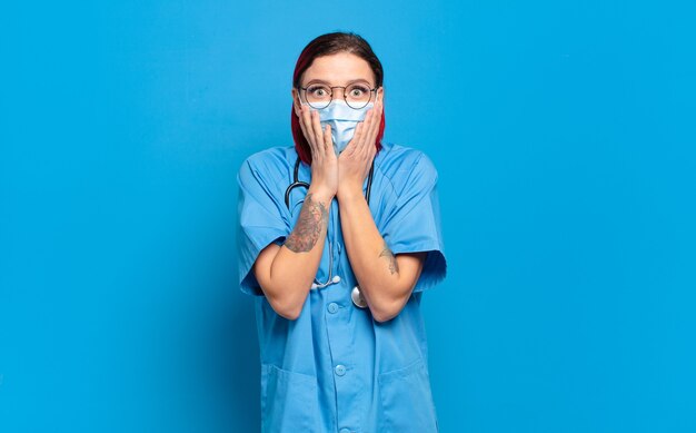 Mujer joven atractiva pelirroja sintiéndose conmocionada y asustada, mirando aterrorizada con la boca abierta y las manos en las mejillas. concepto de enfermera del hospital